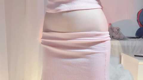 Video of a person in a light pink ribbed dress, pulling it down to expose their buttocks, revealing fair skin. Background includes a white crib, stuffed animals, and a pastel-colored wall.