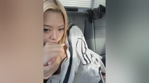 Video of a young Asian woman with blonde hair, wearing a white karate gi, sitting in a car, performing fellatio on a man's erect penis, visible from the side.