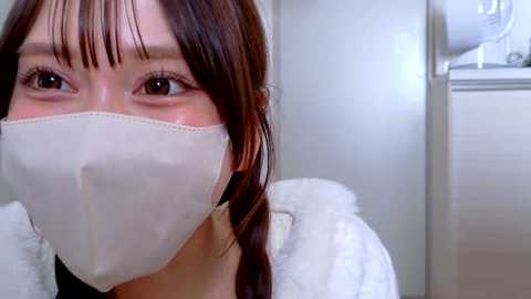 Video of an East Asian woman with light skin and straight brown hair, wearing a white face mask and white bathrobe, in a modern bathroom with tiled walls.