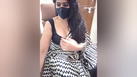 Video of a woman in a black mask, black dress with a black-and-white pattern, sitting on a tan chair in a room with a brown door.