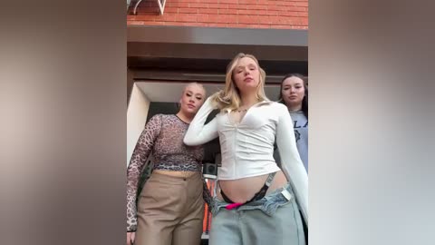 Video of a blonde woman in a white crop top, low-cut jeans, with two women in leopard-print tops behind her, in a modern building with brick walls.