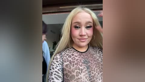 Video of a young woman with long blonde hair, wearing a leopard-print top, applying makeup, in a dimly lit room with blurred figures in the background.