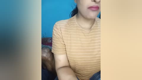 Media: A video of a woman with light skin, wearing a striped beige t-shirt, sitting on a couch. Her lips are slightly parted, and she has dark hair pulled back. The background features a blue wall and a patterned cushion.