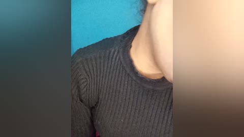 A close-up video of a person's neck and shoulders wearing a dark gray, ribbed knit sweater against a teal and beige background. The texture of the sweater is visible.
