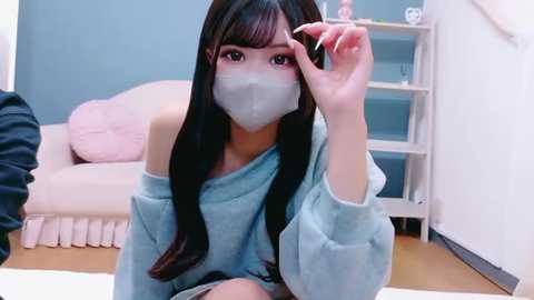 Video of an East Asian woman with long black hair, wearing a light blue off-shoulder sweater, face mask, and makeup, sitting on a white rug in a pastel-colored room.