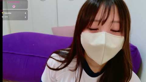 Media: Video of a young Asian woman with long dark hair wearing a white surgical mask, sitting on a purple cushion in a white room.