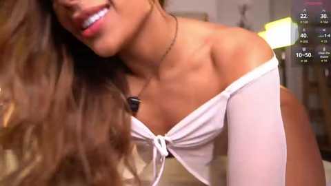 Video of a smiling, light-skinned woman with long brown hair, wearing a white off-shoulder top, in a dimly lit room with a digital timer in the top right corner.