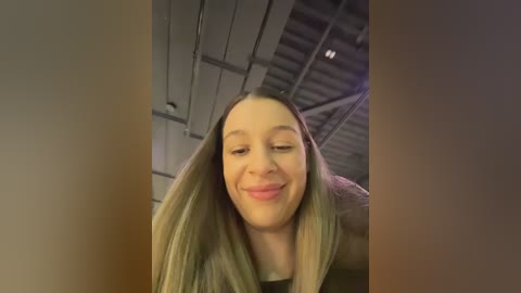 Video of a smiling young woman with long blonde hair, standing in a dimly lit, industrial-style room with exposed beams and metallic structures.