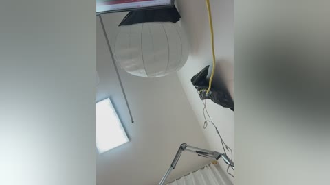 Video of a modern, minimalist room with a white, textured, fabric-covered ottoman against a white wall. A yellow extension cord runs along the wall, and a silver studio light with adjustable arm is in the foreground.