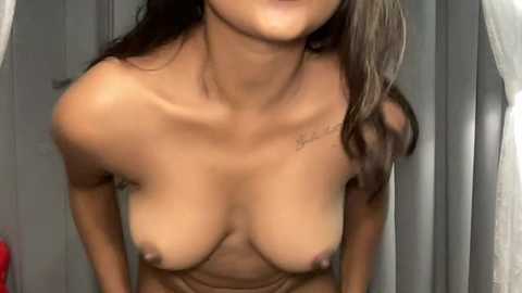 Video of a topless woman with medium-sized breasts, dark hair, and a tattoo on her neck, standing in front of a gray vertical panel, indoors.