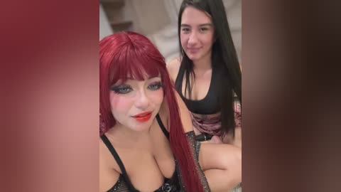 Video of two women, one with red hair and heavy makeup, and the other with long black hair, both wearing revealing black clothing, sitting closely indoors.