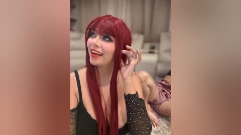 Video of a smiling woman with long red hair and black lace top, posing indoors with blurred background, revealing a living room setting.