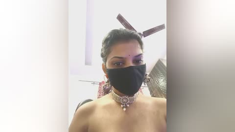 Video of a topless woman with medium skin tone, wearing a black mask, a gold necklace, and a red bindi, standing in a modern room with white walls and a ceiling fan.