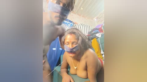Media: Video of a topless woman with medium brown skin, wearing a blue tape over her mouth, lying on a green couch. A man with dark skin, wearing sunglasses, stands behind her.