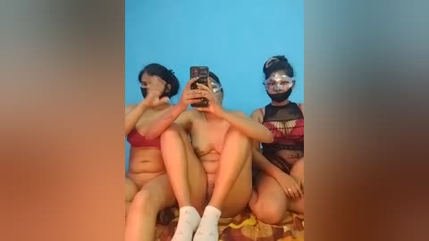 Video of three women, one nude, one in a red bra, and one in a black mesh top, sitting on a patterned blanket against a blue background.