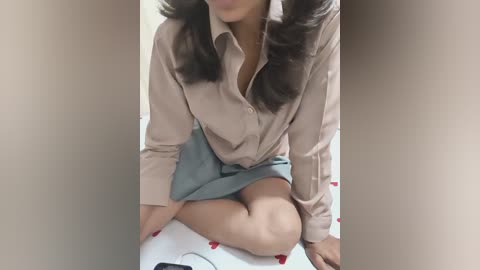 A video of an Asian woman with medium brown skin, wearing a beige blouse and a light blue skirt, sitting cross-legged on a white bedspread with red heart patterns, hands resting on her thighs.