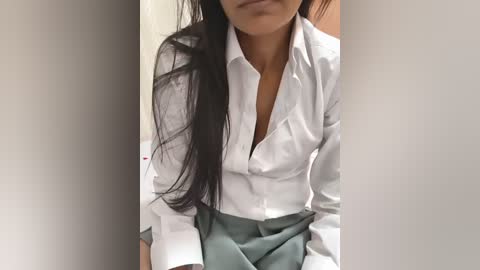 Video of a woman with long black hair, wearing a white button-up shirt and gray skirt, partially unbuttoned, sitting against a beige wall, with a neutral expression.