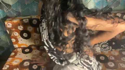 Media: Video of a person with curly black hair, wearing a white and black patterned scarf, lying on a colorful, patterned surface.