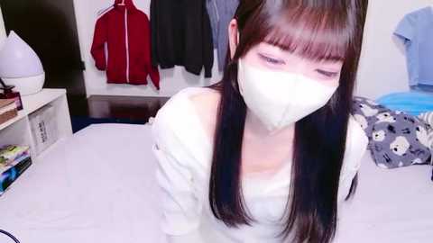 Video of an Asian woman with long black hair, wearing a white face mask and a white top, sitting on a bed in a cozy bedroom with hanging clothes and a white lamp.
