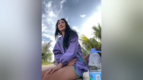 Video of a woman with long black hair, wearing a purple jacket, sitting on a sidewalk, surrounded by palm trees and a blue plastic container.