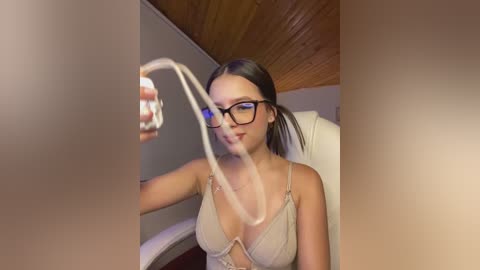 Video of a young woman with straight, dark hair in a ponytail, wearing black-rimmed glasses, a beige bralette, and a necklace, sitting in a dimly lit room with wooden ceiling panels.