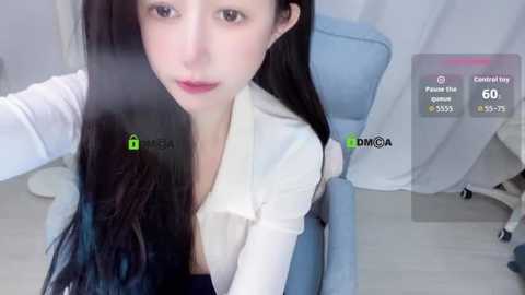 Media: Video of a young Asian woman with long black hair, fair skin, and a slender figure, wearing a white blouse. She is seated in a light blue chair, with a green and white virtual chat interface overlaying the image.