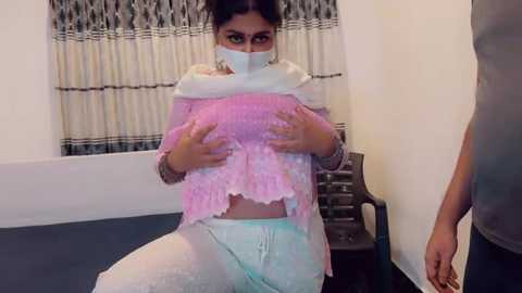 Video of a young woman with medium skin tone, wearing a pink, frilly crop top and white pants, kneeling on a blue carpet, her face partially covered by a white mask.