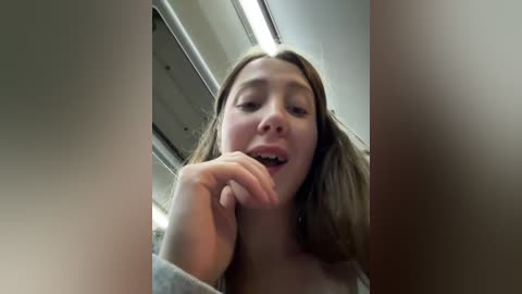 A video of a young woman with fair skin and long, straight brown hair, wearing a gray shirt, slightly smiling and touching her chin. The background shows a dimly lit interior with metallic accents and fluorescent lighting.