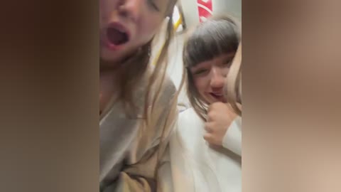 A blurry video of two women in a car, one with long blonde hair, the other with straight dark hair, both wearing light-colored clothing, mouths open, possibly screaming, blurred background.