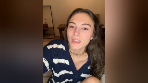 A video of a young woman with long brown hair, wearing a navy-blue striped top, sitting indoors. She has a light complexion and is smiling slightly. Background shows a room with a mirror, wooden furniture, and beige walls.
