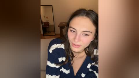 Media: Video of a young woman with fair skin, brown hair, and a delicate facial structure, wearing a navy blue sweater with white wave patterns and a choker necklace. Background features a wooden table, mirror, and beige walls.