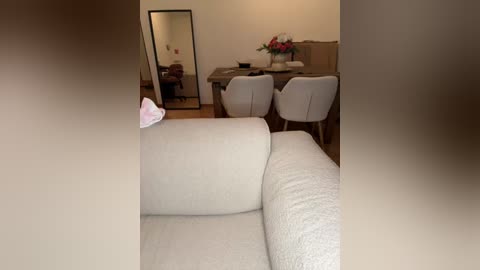 Video of a modern, minimalist living room with beige couch, white chairs, wooden dining table, and mirror.