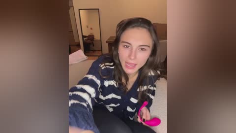 Video of a young woman with long brown hair, wearing a striped sweater and black leggings, sitting on a beige carpet in a cozy, dimly lit room.