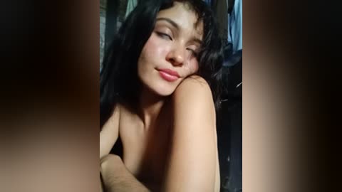 Video of a young, light-skinned woman with curly black hair and closed eyes, resting her head on her arm. Background is blurred, suggesting an indoor setting.