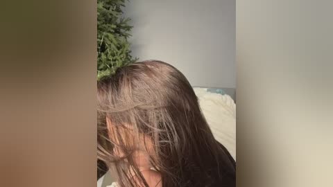 Video of a woman with long, straight brown hair, partially covering her face, sitting on a white bed in a room with light gray walls and a green plant in the background.
