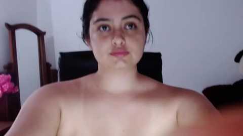 A video of a topless woman with fair skin and dark hair, sitting on a bed in a dimly lit room. The background includes a wooden mirror and a pink flower arrangement.