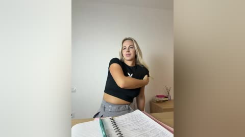 Video of a blonde woman with medium build, wearing a black crop top and gray pants, sitting at a desk with a spiral notebook, in a minimalist room with beige walls.