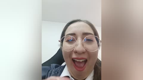 Video of a young Asian woman with light skin and glasses, mouth open in a wide, excited smile, standing in a white-walled room.