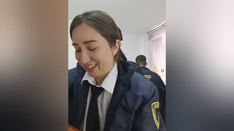 Video of a smiling, light-skinned young woman with long brown hair, wearing a blue police uniform, standing in a classroom with white walls and blinds.