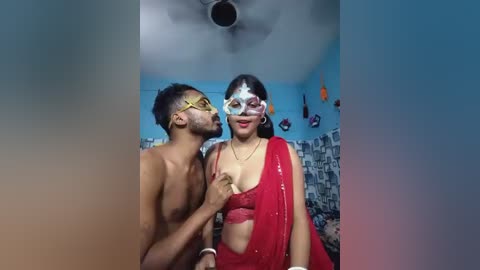 Video of a shirtless man with a mustache wearing a yellow mask, kissing a woman in a red saree and mask in a colorful room with cityscape wallpaper.