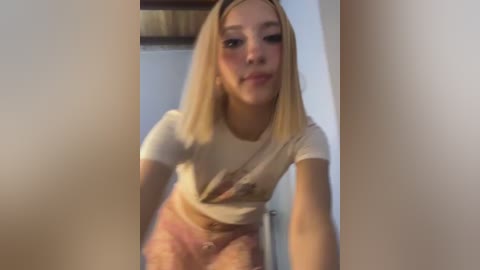 Media: Video of a young woman with straight, shoulder-length blonde hair, wearing a white crop top and pink shorts, standing indoors near a door.