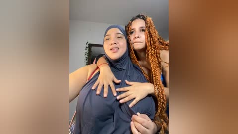 Media: Video of two women, one with a dark blue hijab and the other with orange braids, hugging closely, smiling, in a dimly lit room with blurred background.
