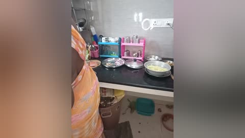 Media: Video of a cluttered kitchen countertop with various cooking utensils and ingredients, including a colorful apron, pink and blue storage shelves, and a white wall.