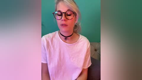 A video of a blonde woman with glasses, wearing a white t-shirt and black choker, sitting against a teal wall, eyes closed, appearing thoughtful.