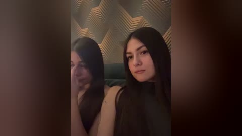 Video of a young woman with long dark hair, wearing a black top, seated indoors with a geometric-patterned wallpaper in the background.