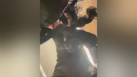 Video of a woman in a black hoodie, crouched in a dark, dimly lit room, fists clenched, looking aggressive.