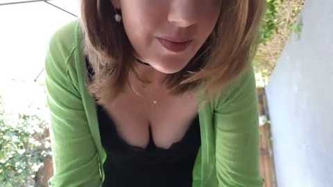 Video of a Caucasian woman with light skin, shoulder-length brown hair, and a green cardigan over a low-cut black top, showing cleavage, in an outdoor setting with blurred trees and a light blue wall.