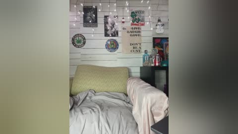 Video of a cozy dorm room with white walls, string lights, a bed with light gray bedding, a green pillow, and various posters.