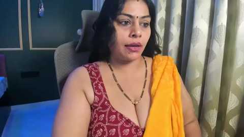 Video of a South Asian woman with medium brown skin, long black hair, wearing a red blouse with a gold necklace, and a yellow sari. She sits on a beige chair in a dimly lit room with green curtains.