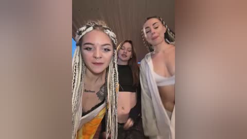 Video of three women in a dimly lit room: one with dreadlocks, one with a tattoo, and one with a white shirt, all smiling.